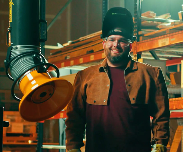 Welder wearing PPE standing next to a wall-mounted fume extractor arm in a workshop, illustrating safe welding practices.